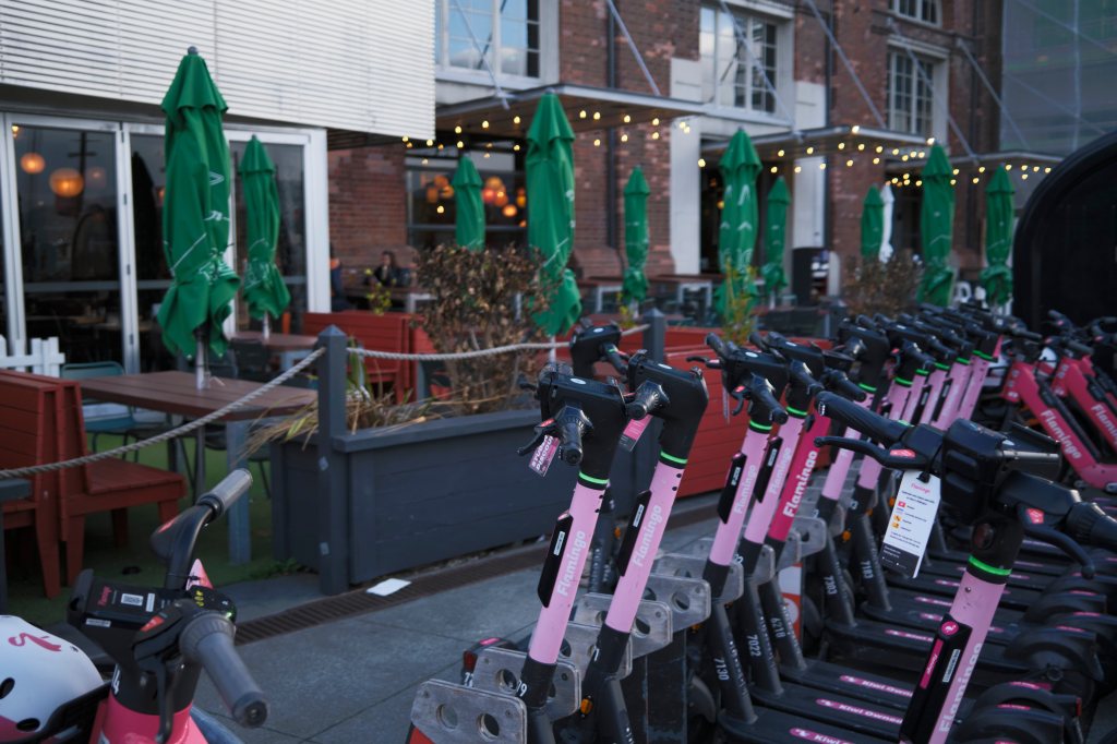 A row of pink electric scooters parked in front of a cafe with green umbrellas and outdoor seating.