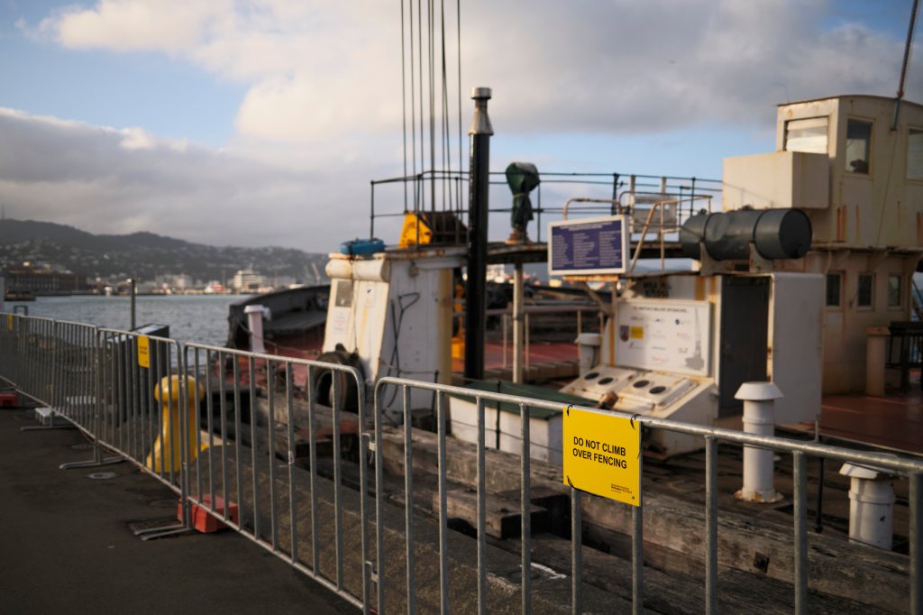 View of a harbor area featuring a fenced-off section with a warning sign that says 'DO NOT CLIMB OVER FENCING,' showing a moored boat and distant cityscape in the background.