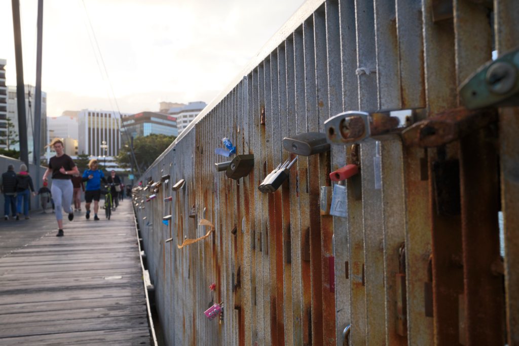 Close-up of a weathered bridge railing adorned with various padlocks, while joggers and cyclists pass by in the background.