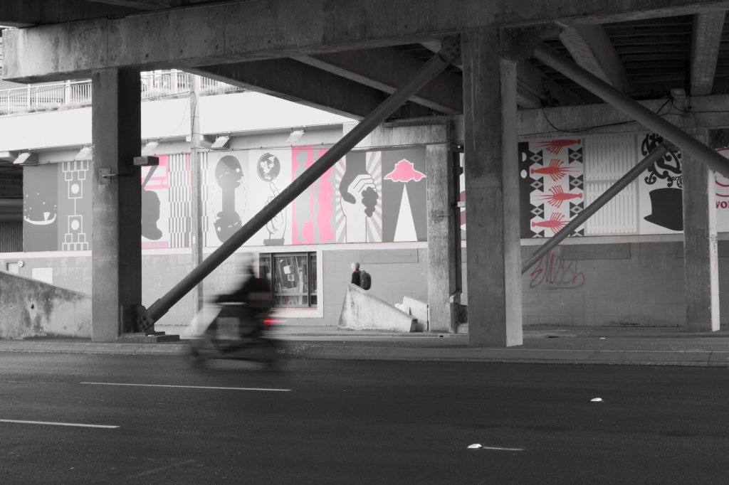 A blurred motorcycle passes under a bridge featuring colorful murals and concrete pillars.