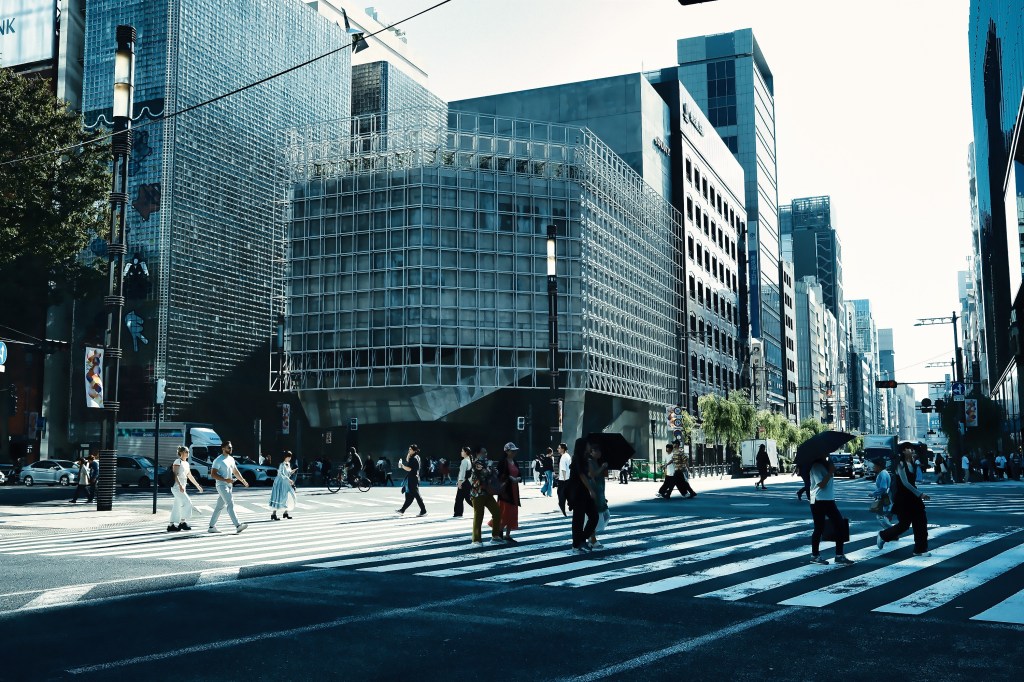 Busy urban street with pedestrians crossing at a zebra cross, surrounded by modern buildings and clear blue sky.