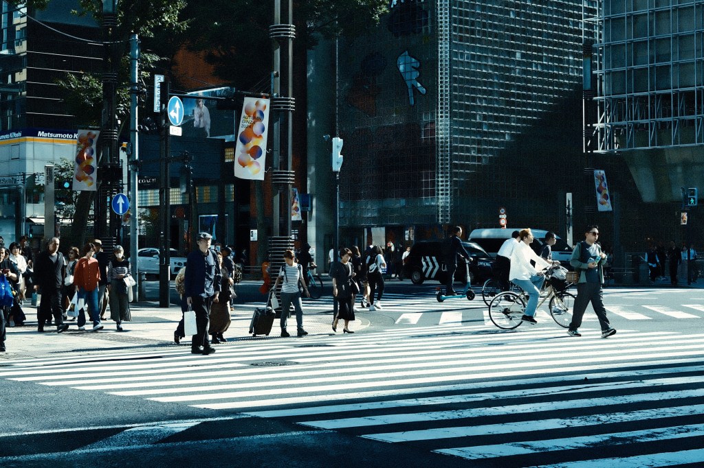 Busy urban crosswalk with pedestrians and a cyclist, modern buildings in the background, and traffic signs visible.