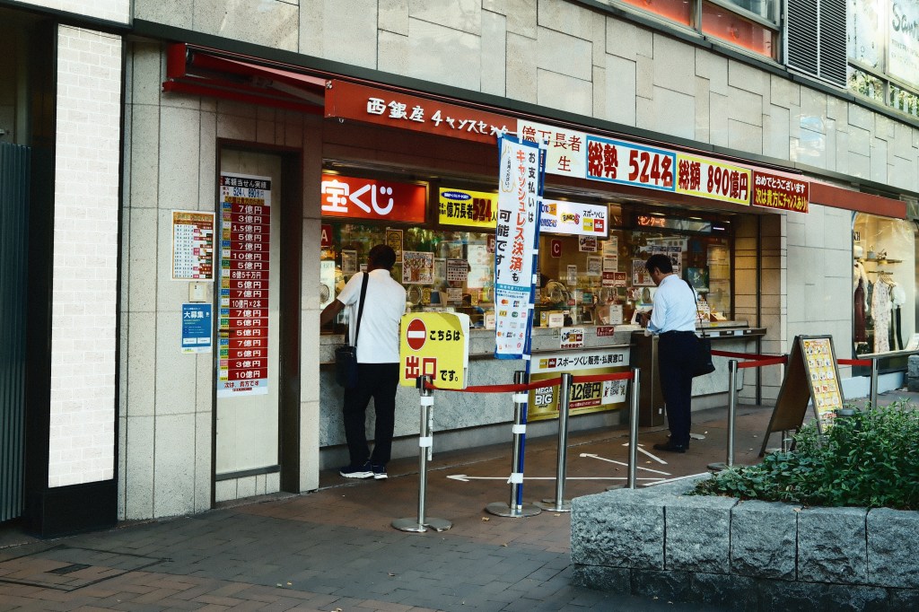 A lottery ticket booth in an urban setting, with two people interacting with the service counter. The booth displays various lottery options and prizes on the windows.