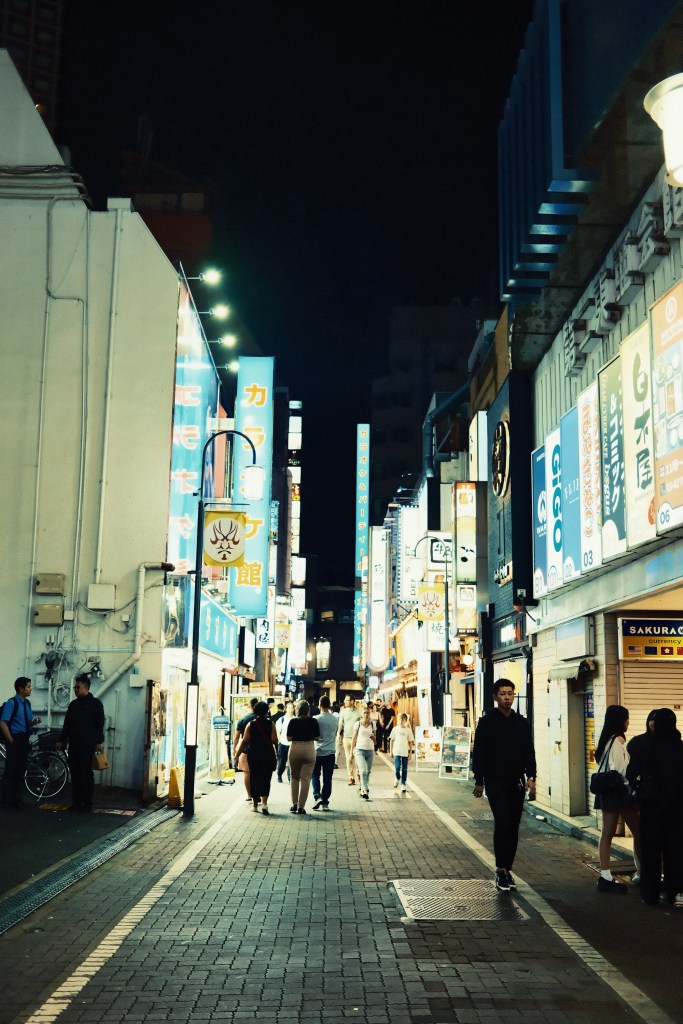 A bustling urban street at night, lined with colorful neon signs and people walking along the paved pathway.