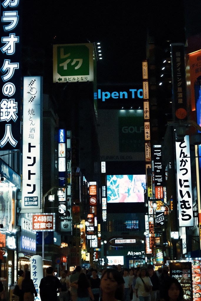 A bustling city street at night filled with bright neon signs and advertisements, showcasing various restaurants and businesses, with a crowd of pedestrians walking along the sidewalk.