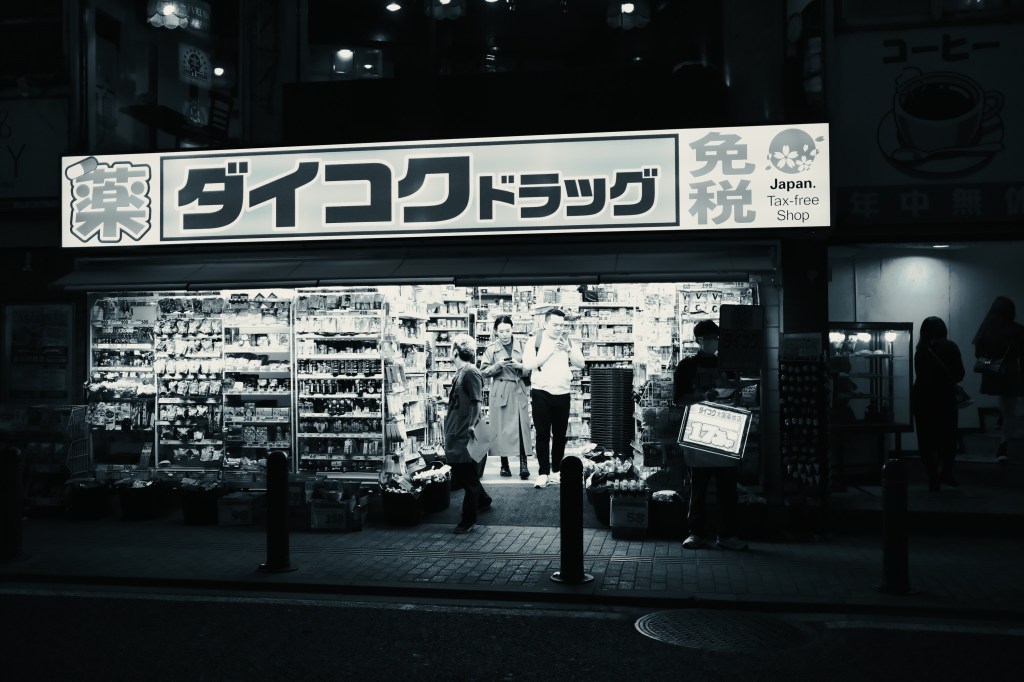 A brightly lit pharmacy storefront in Japan, displaying a variety of products. Two people are conversing in front of the store, which has a tax-free sign and is decorated with Japanese characters.