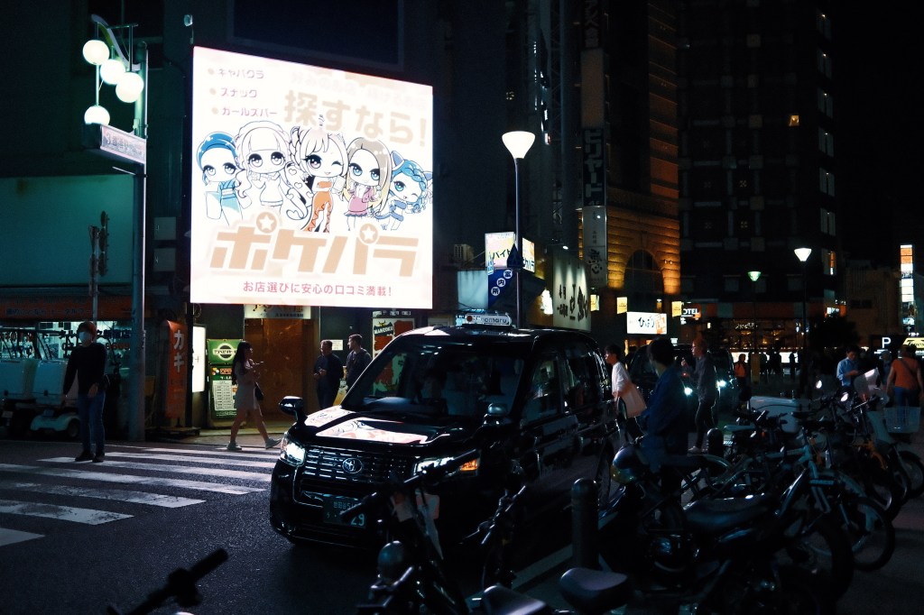 A busy street scene at night featuring a black taxi in the foreground and pedestrians crossing the road. A large illuminated billboard displays colorful animated characters alongside Japanese text.