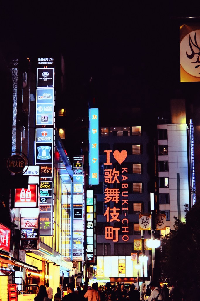 A vibrant street scene in Kabukichō at night, illuminated by colorful neon signs and advertisements, showcasing various businesses.