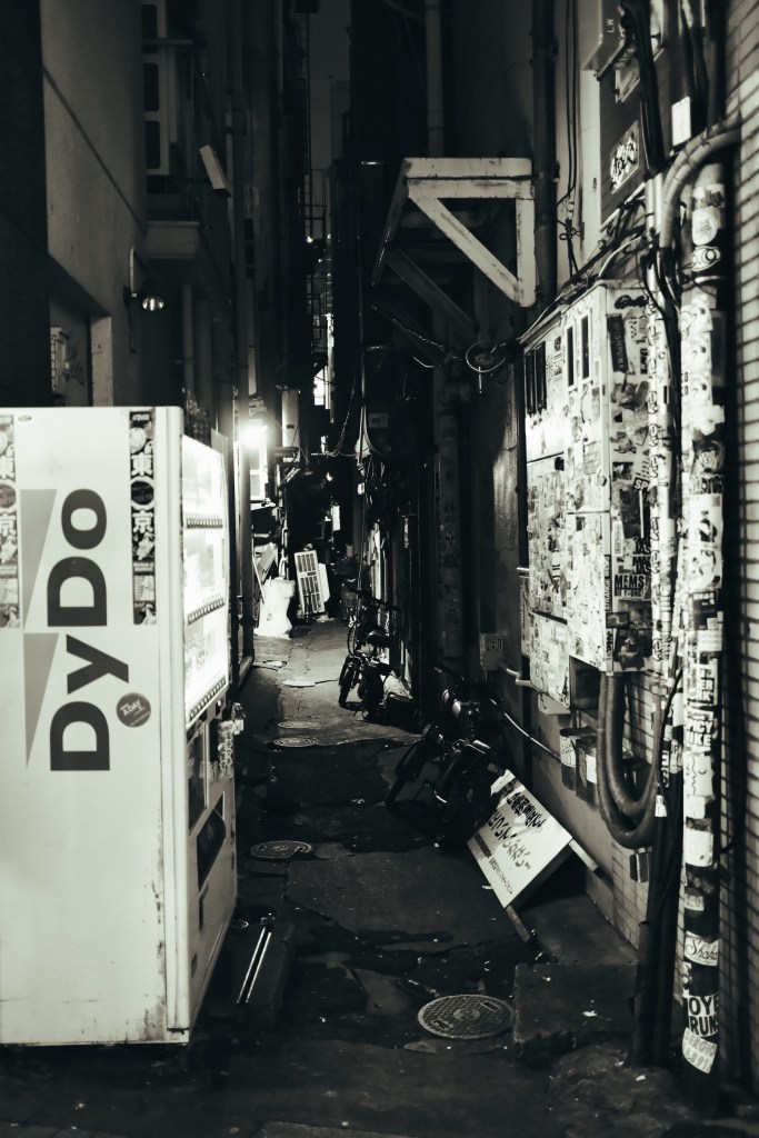 Narrow alleyway at night with a vending machine on the left, surrounded by buildings featuring graffiti and posters.