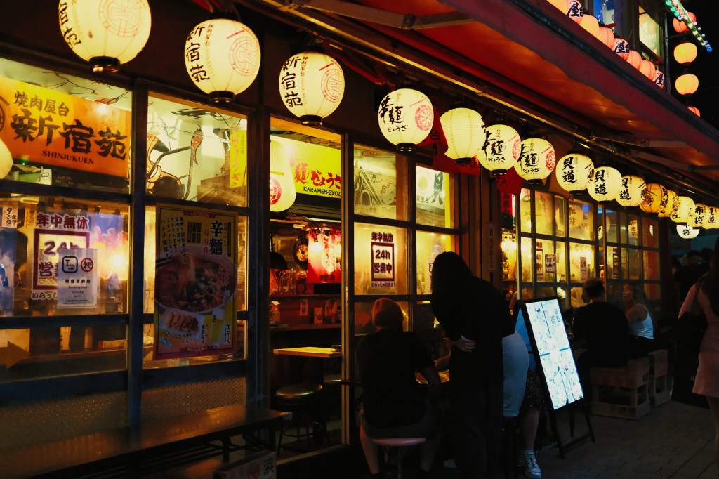 A nighttime view of a bustling restaurant storefront adorned with colorful lanterns, showcasing a variety of signs and menus, with customers seated outside and inside.
