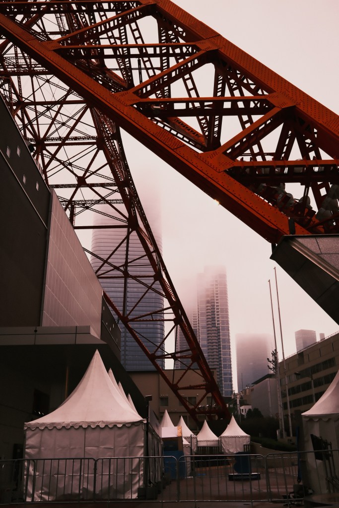 A view of the Tokyo Tower's red steel structure against a backdrop of fog and modern buildings, with white tents in the foreground.