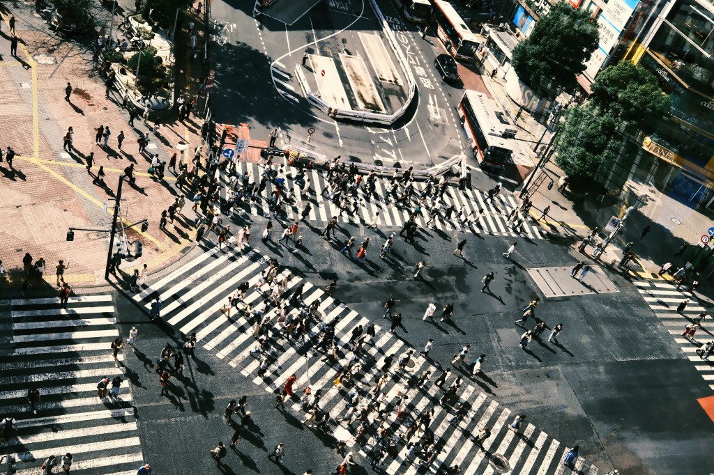 Aerial view of a busy intersection with numerous people crossing pedestrian zebra stripes on a sunny day.