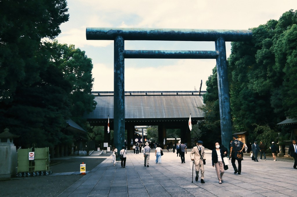 View of a large torii gate at the entrance of a traditional Japanese shrine, with people walking along a stone pathway surrounded by greenery.