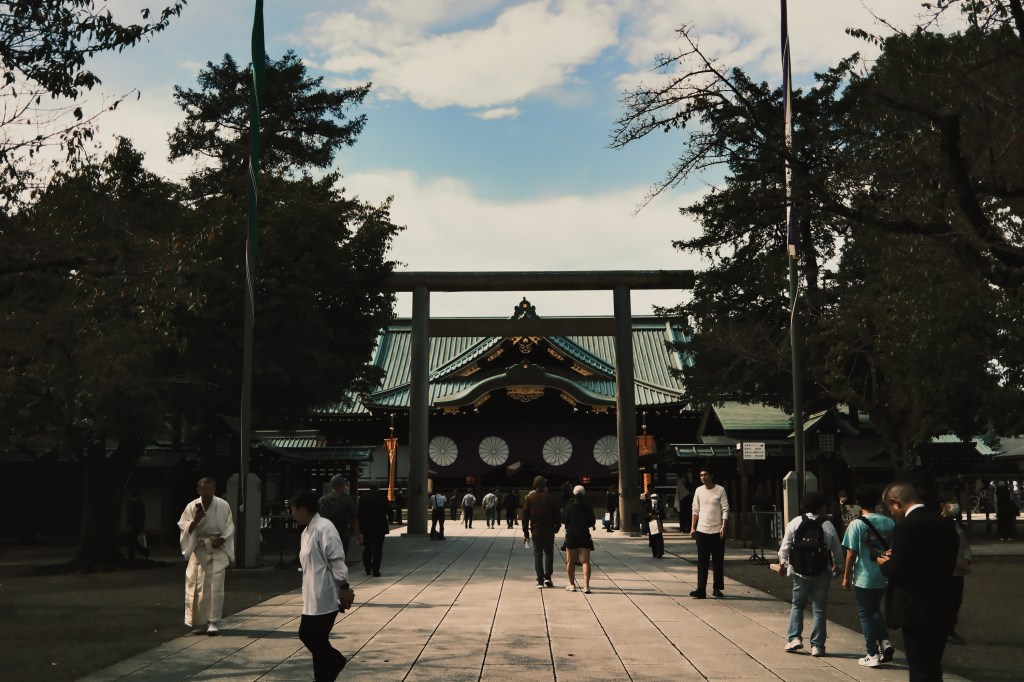 A pathway leading to a traditional Japanese shrine, with people walking towards the entrance under a large torii gate, surrounded by trees and blue sky.
