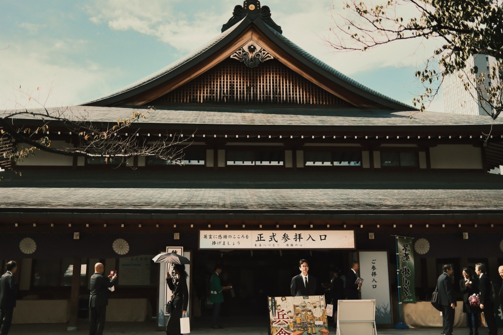 Entrance of a traditional Japanese building with a slanted roof and wooden architecture, featuring a sign for formal entry. People dressed in formal attire gathered outside.