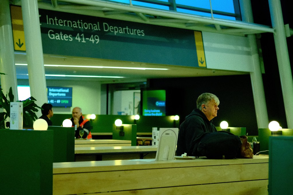 Interior of an airport with a sign indicating  International Departures Gates 41-49. Two people are engaged in conversation at a table, while another person sits alone nearby, looking towards the camera. Soft lighting is provided by round lamps on the tables.