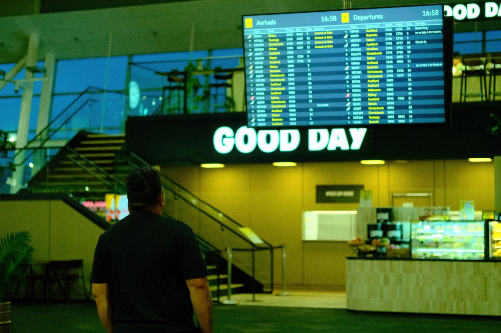 A person looking up at a digital flight information board in an airport, displaying arrivals and departures. The background features a café with seating and a sign that says 'GOOD DAY'.