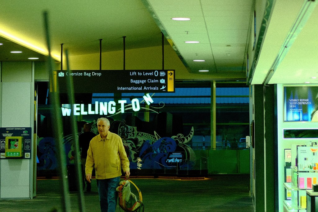 An older man walks through an airport terminal, carrying a bag, with signs indicating 'Baggage Claim' and 'International Arrivals' in the background, and 'Wellington' displayed prominently.