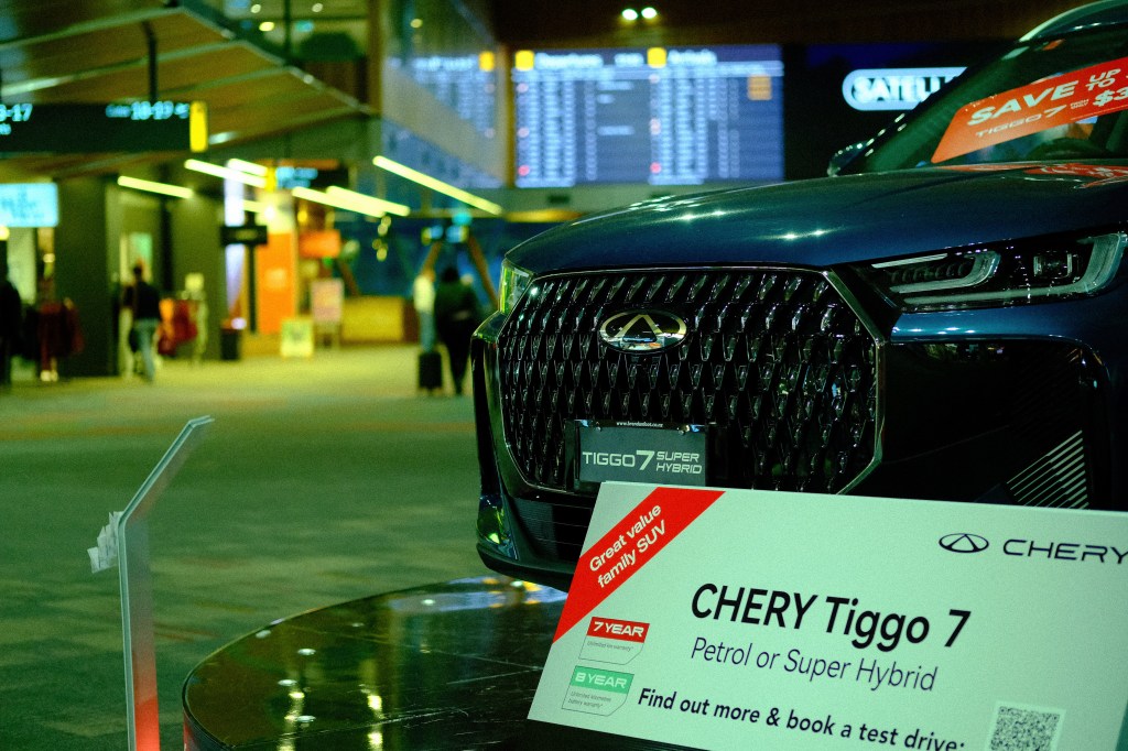 Close-up of a Chery Tiggo 7 SUV displayed in an airport terminal, featuring a prominent grille and promotional signage highlighting its petrol and hybrid options.