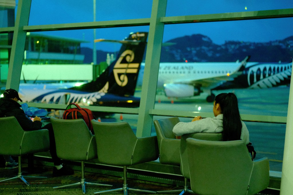 Interior of an airport terminal at dusk, showing two passengers seated in modern chairs, with a view of an airplane through large windows.