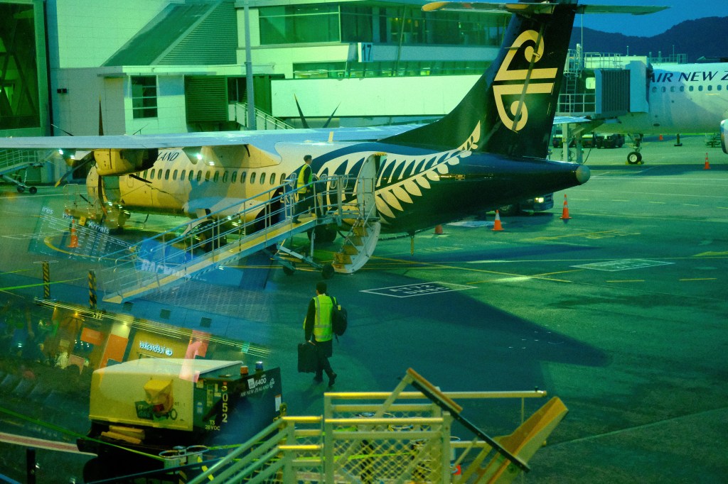 A view of an Air New Zealand regional aircraft at an airport, with a passenger boarding ramp and ground crew in view. The scene is illuminated with evening lighting.