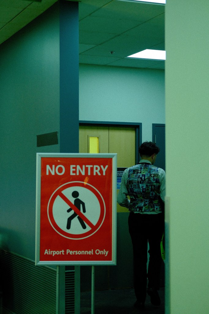 A man wearing a patterned vest is seen walking away from a doorway, next to a bright red sign that reads 'No Entry - Airport Personnel Only'.