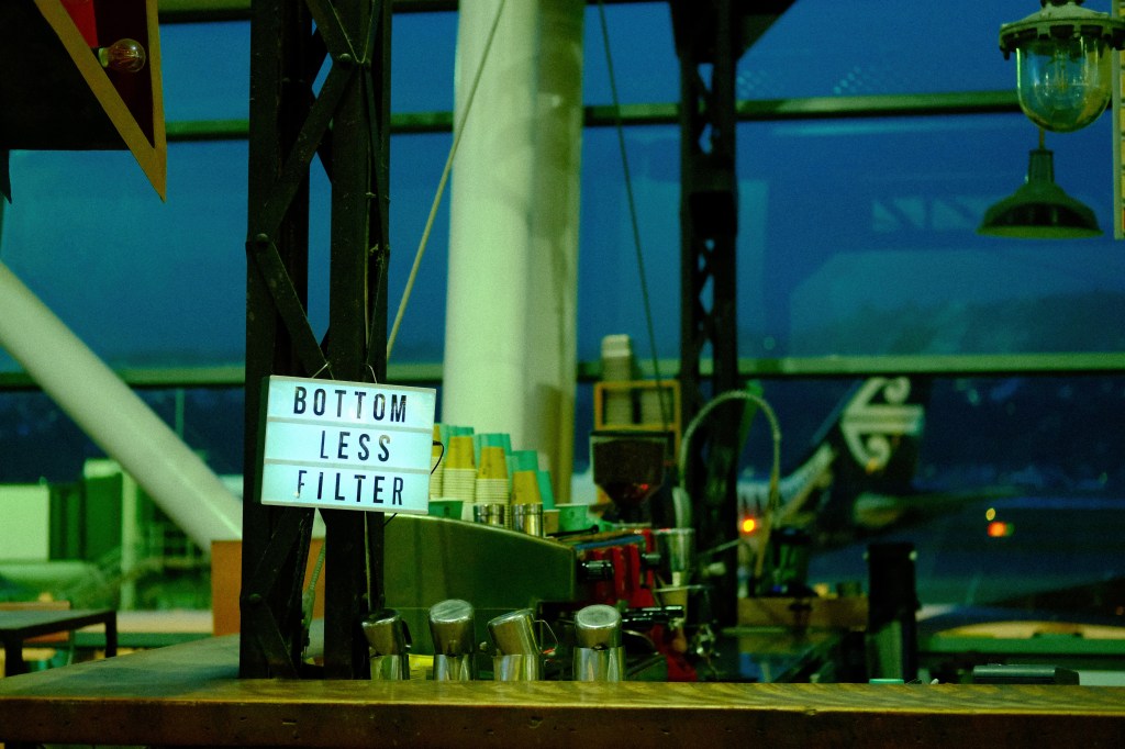 A coffee shop counter featuring a lightbox sign that reads 'BOTTOM LESS FILTER'. The background includes coffee equipment and colorful cups, with an airport runway visible through large windows.