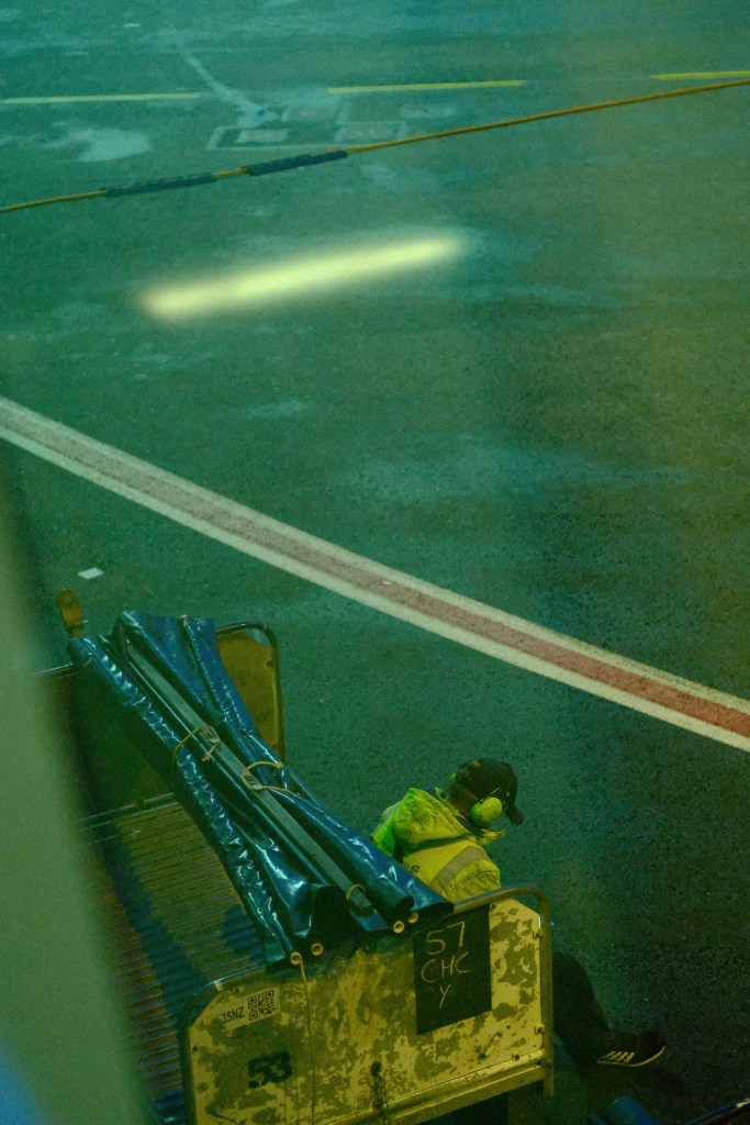 A ground crew member wearing a high-visibility jacket and headphones sits on a luggage cart at an airport, with a wet, illuminated runway in the background.