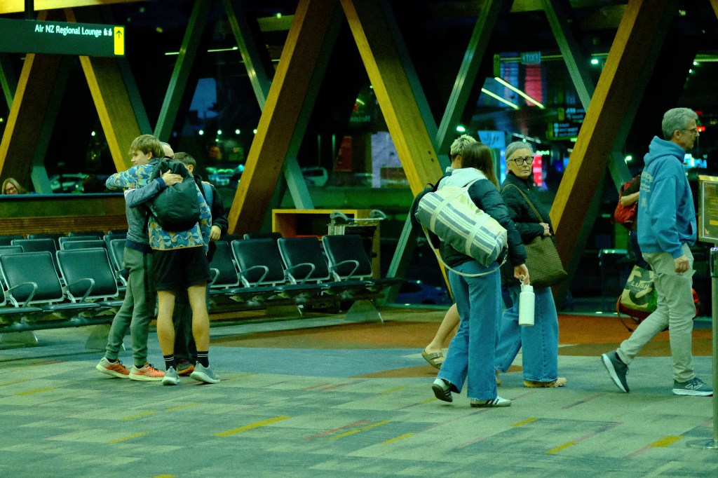 People interacting in a vibrant airport setting during the evening, with some embracing and others walking by, surrounded by seating areas and various signs.