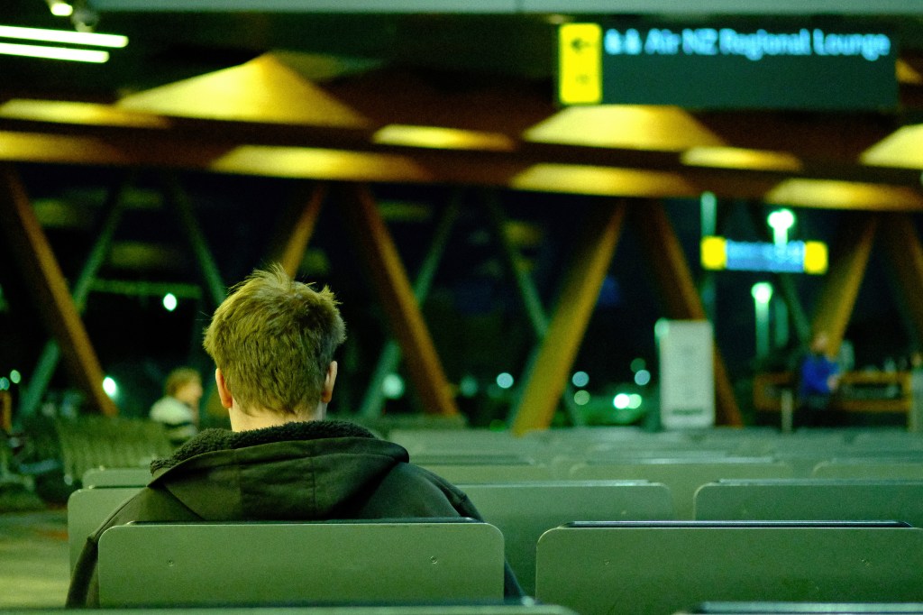 A person sitting alone in an airport lounge at night, viewed from behind, with empty seating in the foreground and a sign indicating the Air New Zealand Regional Lounge in the background.