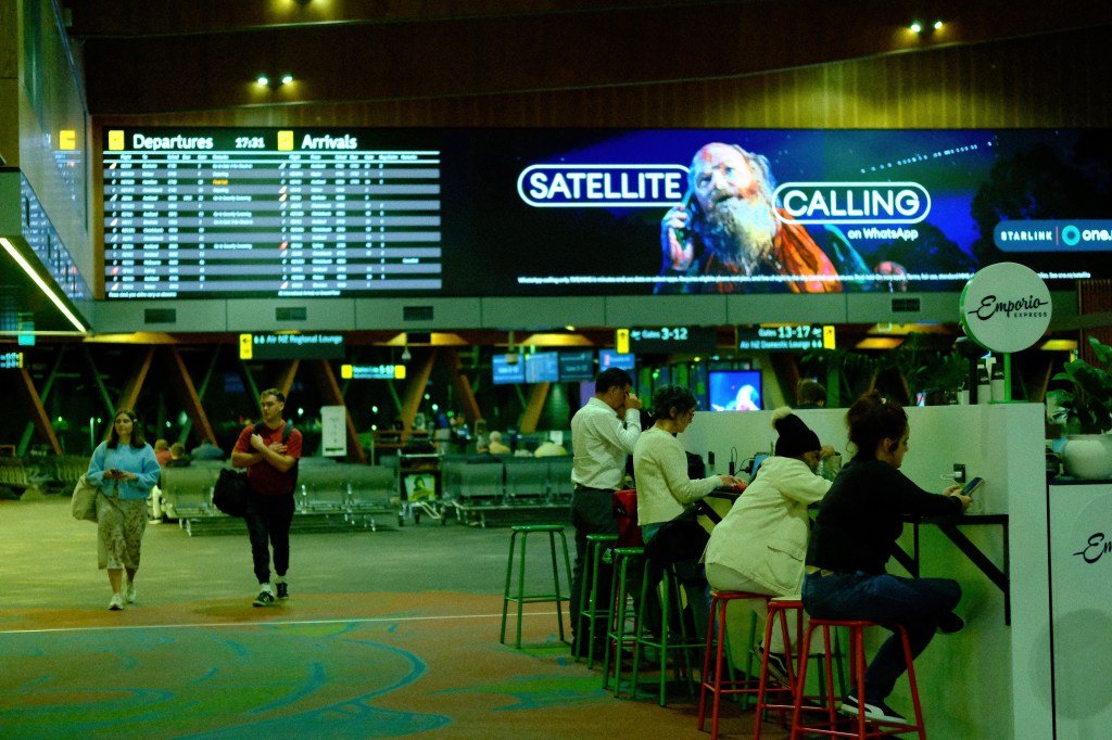 Interior of an airport terminal featuring a digital departures and arrivals board, a large advertisement for satellite calling, and travelers sitting at a café.