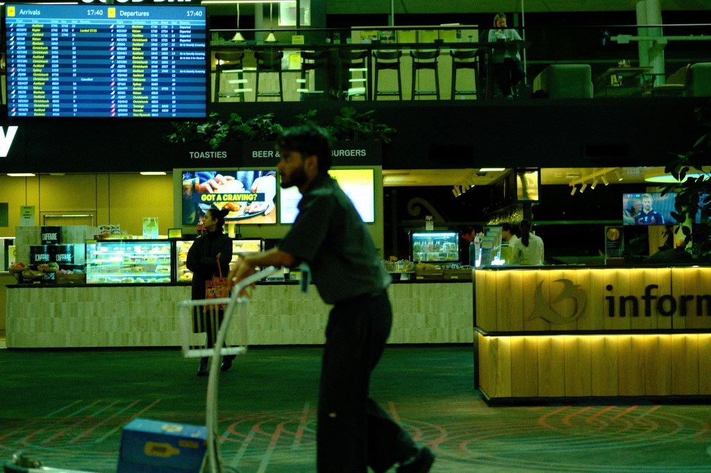 An airport terminal interior showing a flight information board, a food counter, and travelers, with one person pushing a luggage cart in the foreground.