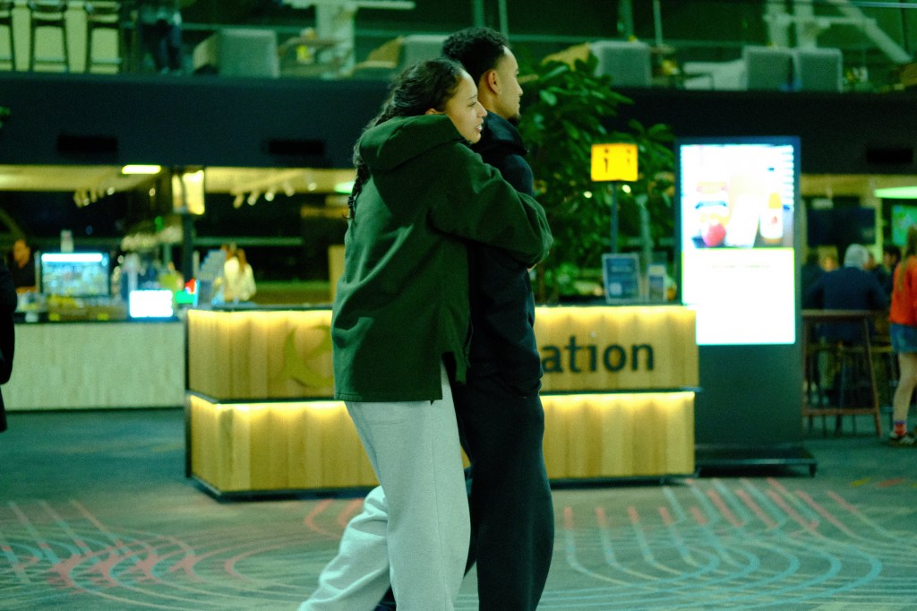 A couple embracing in a dimly lit indoor space, with a coffee shop and digital screens in the background.
