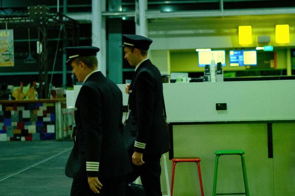 Two airline pilots in uniform walking through an airport terminal, with a bar area and seating in the background.