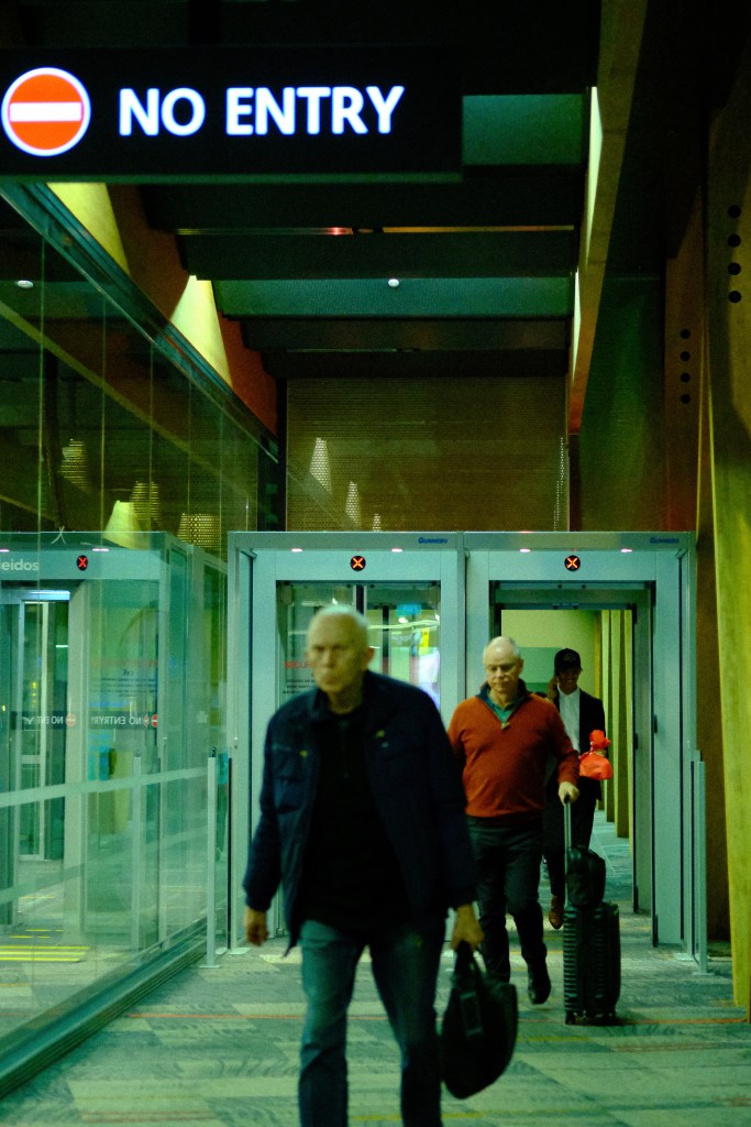 A group of travelers walking through an airport corridor with a 'No Entry' sign visible above them.