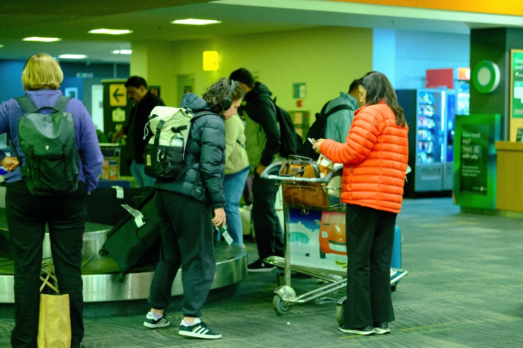 Passengers waiting at the baggage claim area in an airport, some checking their luggage on a cart.