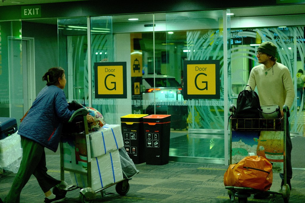 An elderly woman pushing a luggage cart at an airport entrance labeled 'Door G', while a young man stands nearby with his own cart.