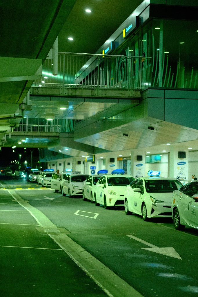 Line of white taxis parked at an airport at night, with a modern terminal building in the background.