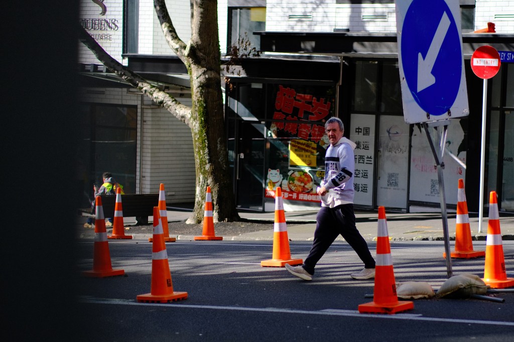 A man walking on a street surrounded by traffic cones, with a sign indicating the direction to turn. Shops are visible in the background.