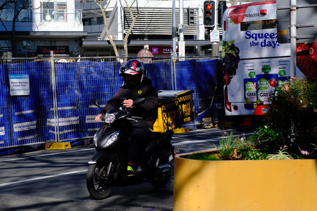 A person wearing a helmet riding a scooter along a city street near construction barriers and a bus advertising fresh juice products.