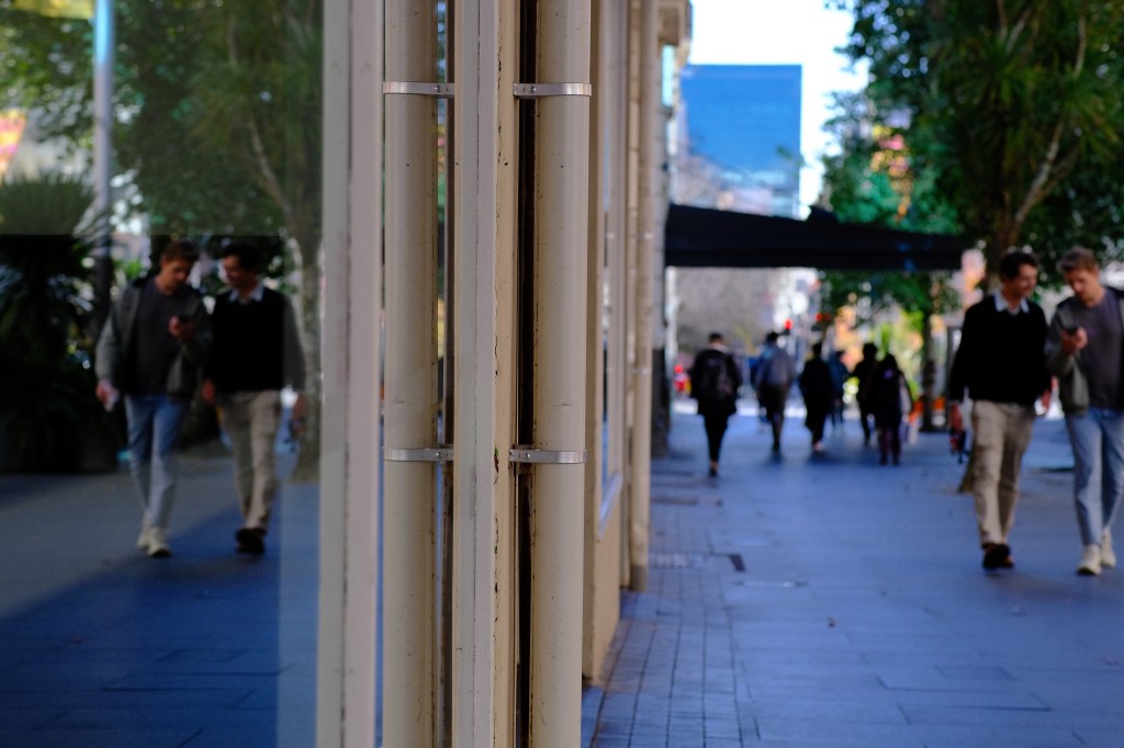 Two people walking side by side on a city street, with reflections visible in a glass window. In the background, other pedestrians are moving along the sidewalk, surrounded by greenery and urban architecture.