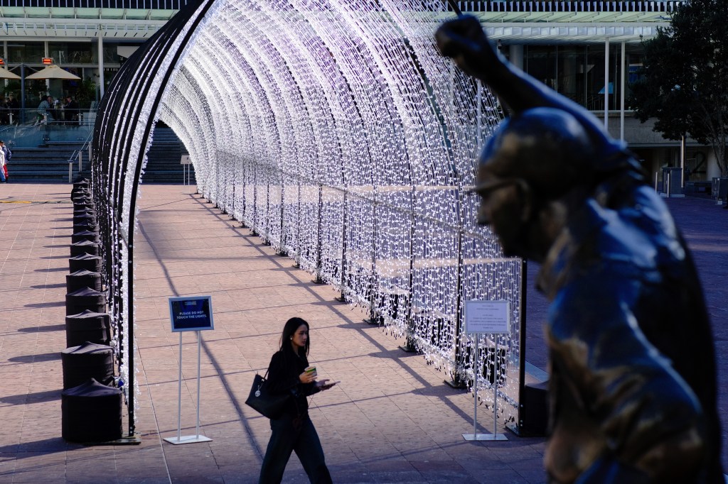 A woman walks past an illuminated archway made of lights in an outdoor plaza, with a bronze statue visible in the foreground.