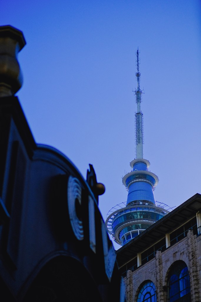 View of a tall communications tower against a clear blue sky, with buildings partially visible in the foreground.