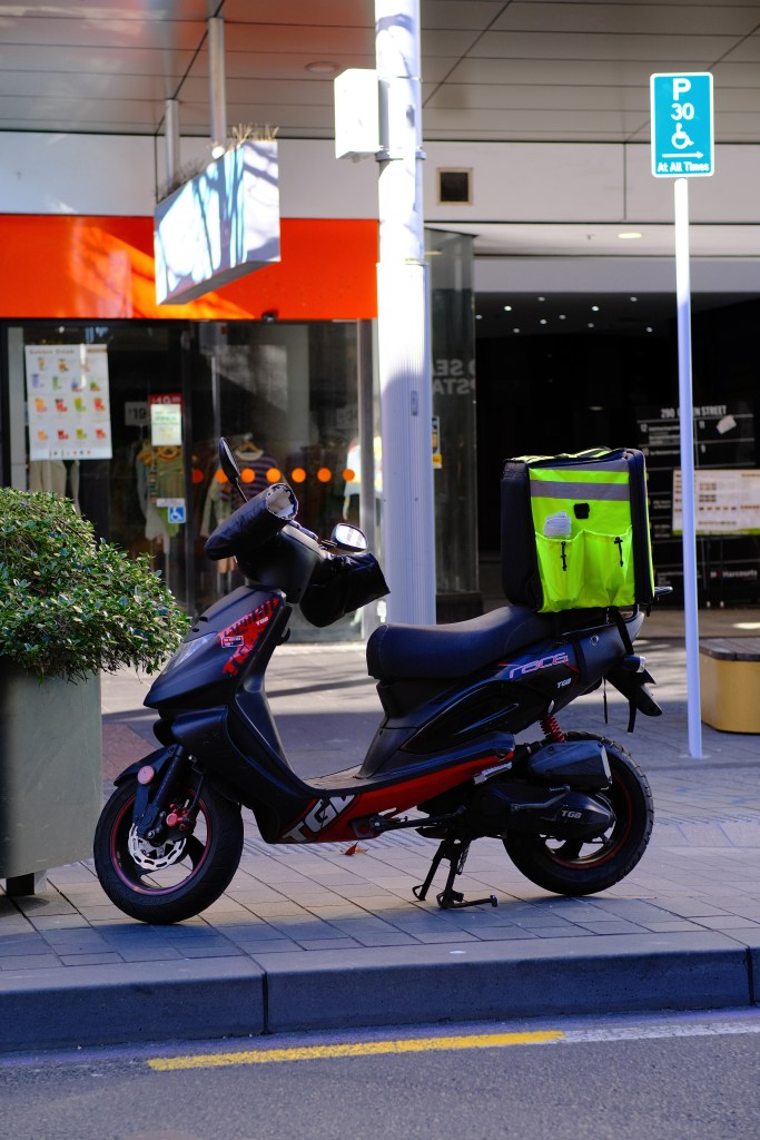 A black and red scooter parked on a sidewalk with a bright yellow delivery bag on the back. In the background, there's a building facade with signs and a parking sign.