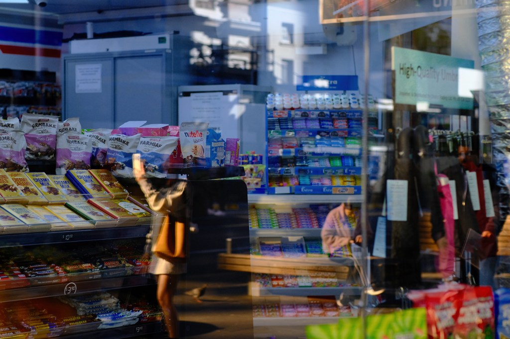 A reflection of a convenience store with shelves stocked with snacks, candies, and beverages. A person is seen through the glass, holding a phone, while another customer browses the store.