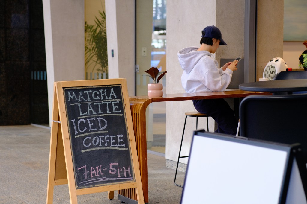 A chalkboard sign advertising matcha lattes and iced coffee with business hours from 7 AM to 5 PM, positioned in a café setting where a person in a hoodie is sitting at a wooden table, using a smartphone.