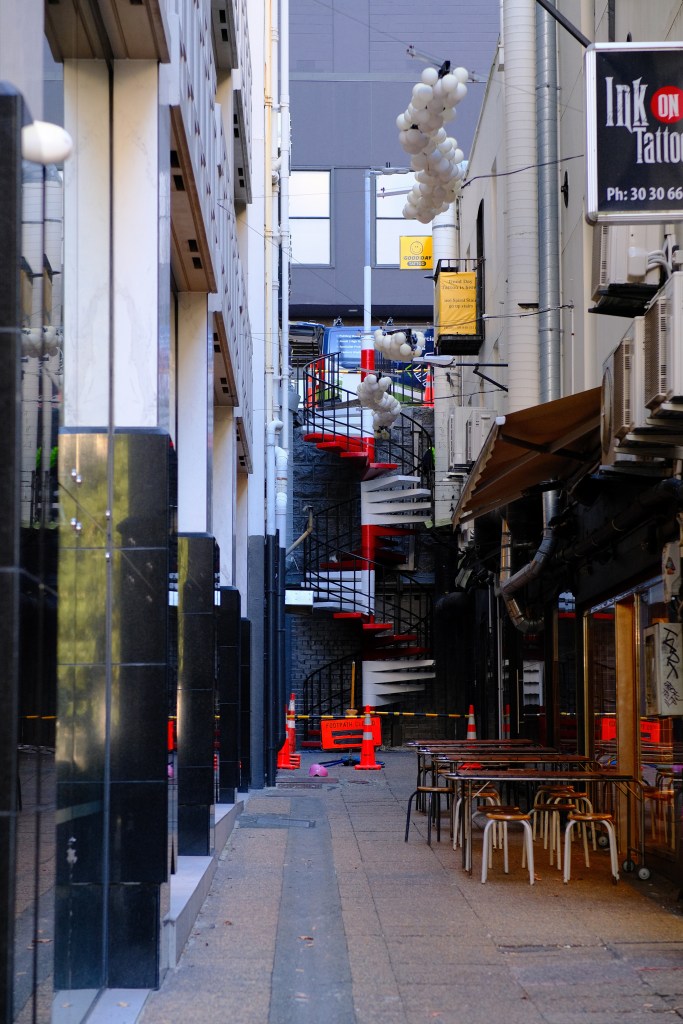 Narrow alleyway featuring a spiral staircase adorned with balloons, surrounded by buildings, reflective glass surfaces, and outdoor seating areas.