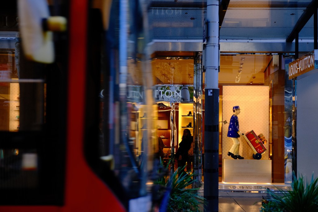 A storefront of a Louis Vuitton store, featuring a colorful mannequin wearing a blue uniform and pushing a red suitcase, with blurred reflections of a bus in the foreground.