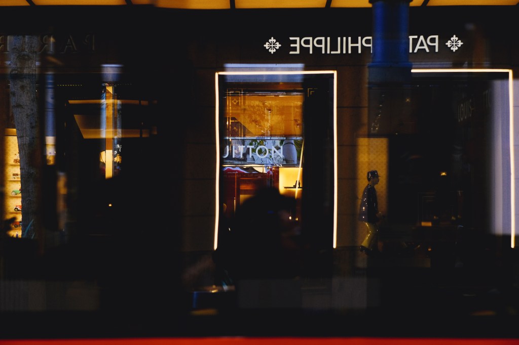 Reflected view of luxury storefronts with a mannequin and illuminated signage, showcasing brands in a high-end shopping district.
