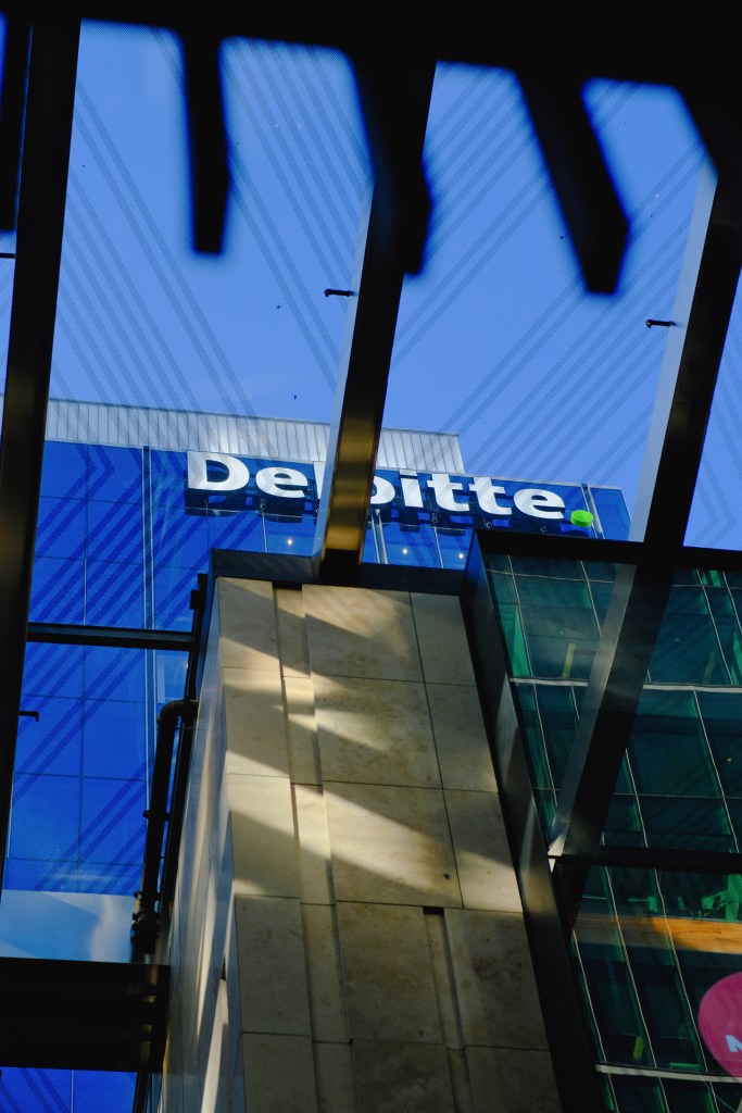 View of a Deloitte building with a blue sky in the background, captured from below through a glass structure.